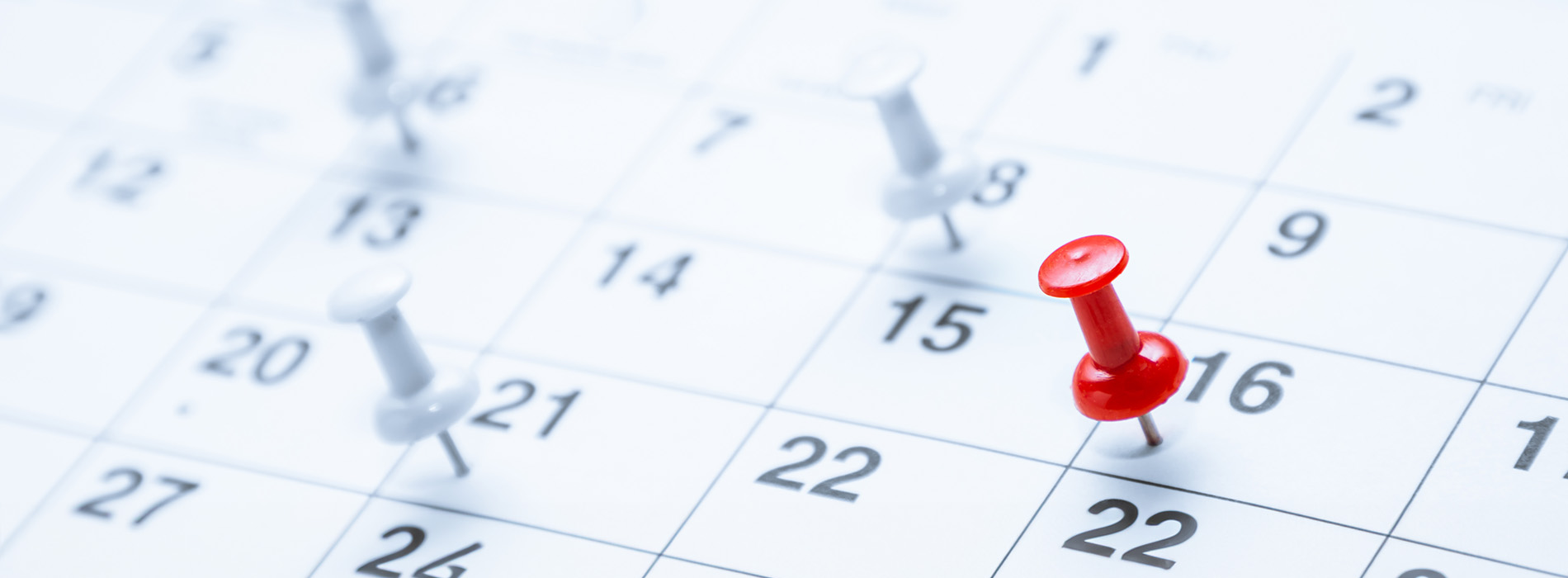 The image depicts a close-up view of a calendar with a red thumbtack on it, placed over a section of dates showing the month of April with some days highlighted.