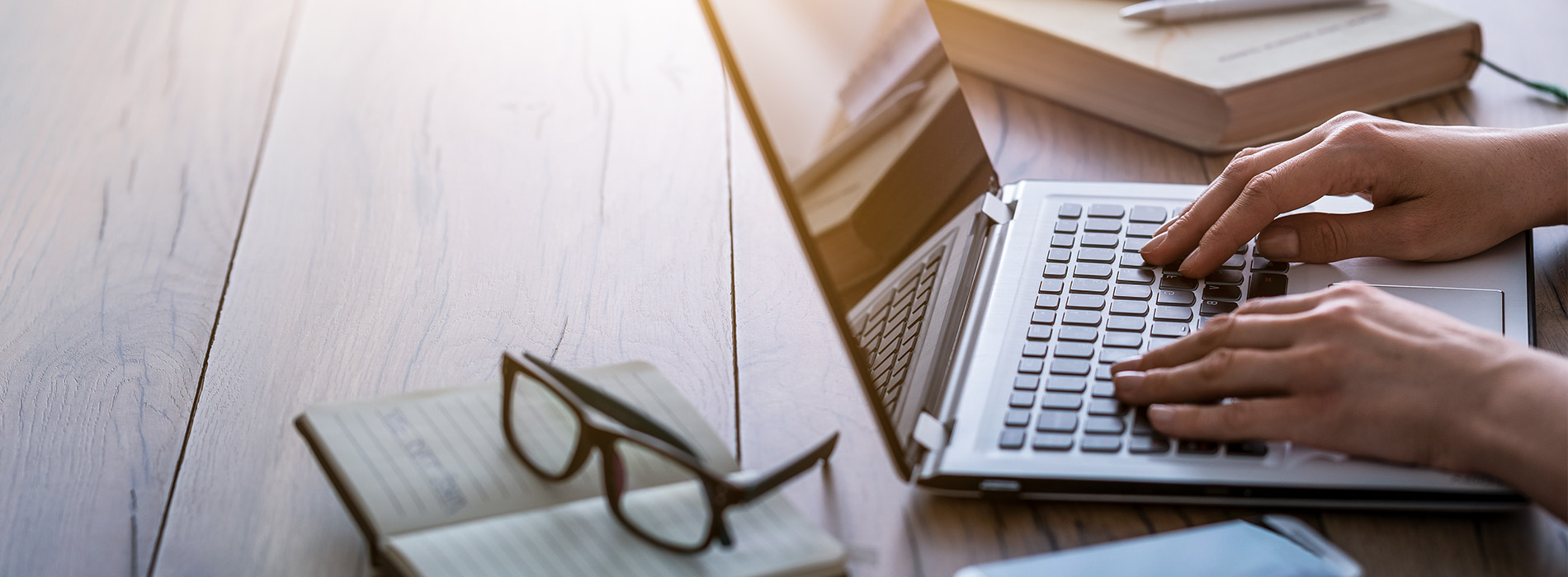 A person working on a laptop at a desk with books and a pen.
