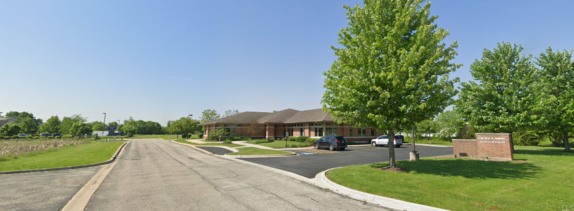 The image shows a daytime scene of a residential street corner with a clear sky, a tree-lined sidewalk, a brick building with a sign, and a paved parking lot.