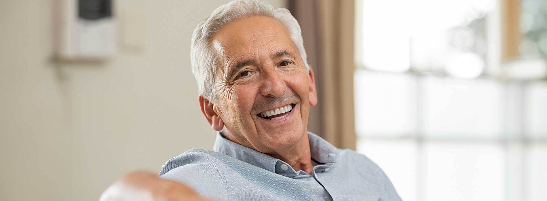 An older man with gray hair is sitting comfortably with his legs crossed, smiling slightly at the camera.