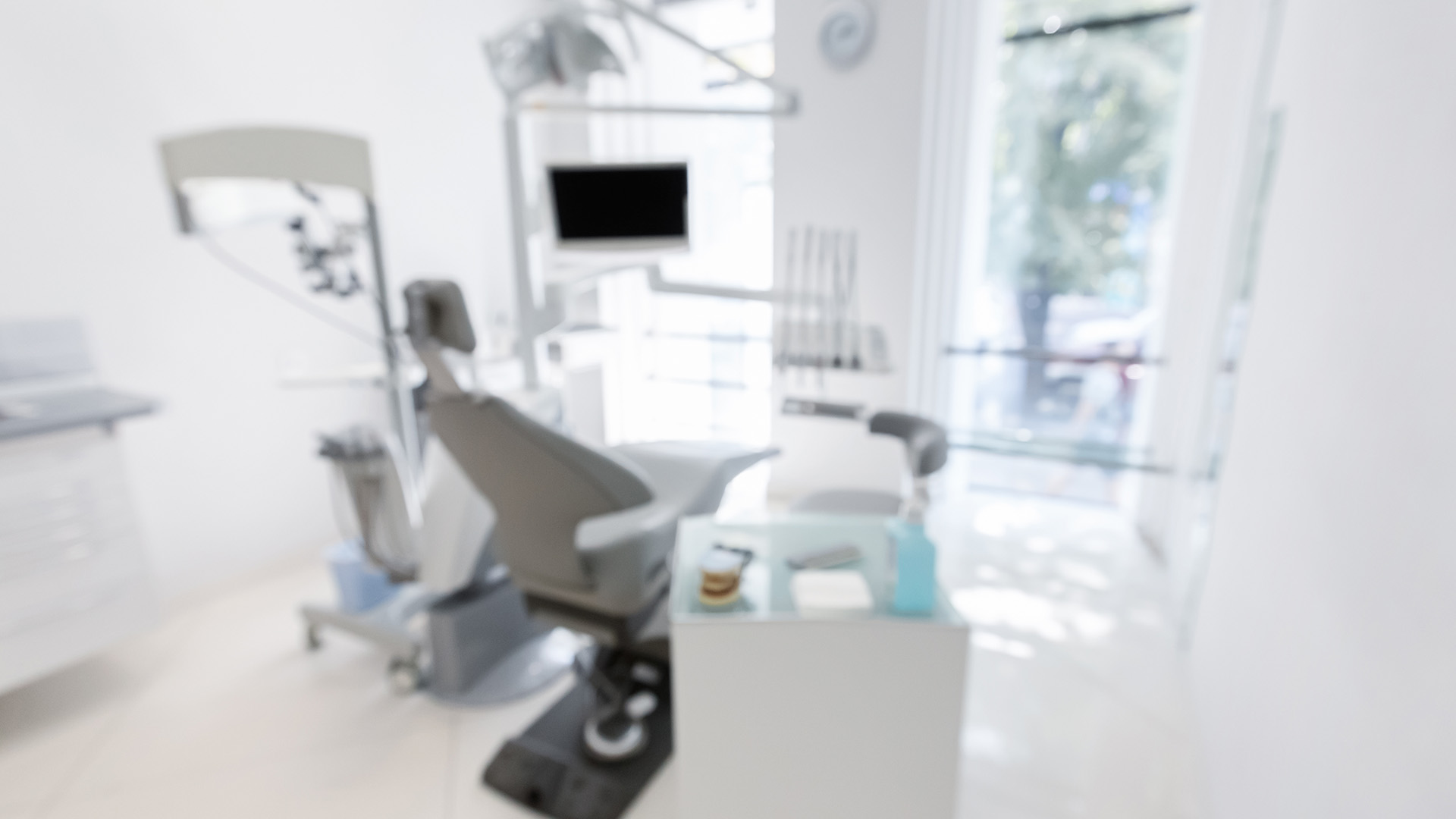 The image shows an interior view of a dental office with a modern chair and equipment visible, set against a brightly lit room with a large window allowing natural light to enter.