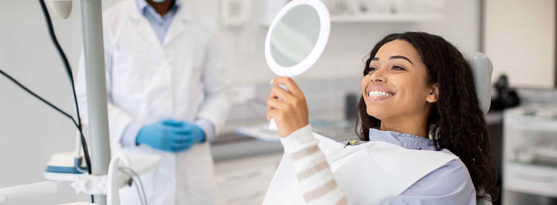 A woman in a dental chair being attended to by a dentist, with a smiling face and a mirror reflecting her cheerful expression.