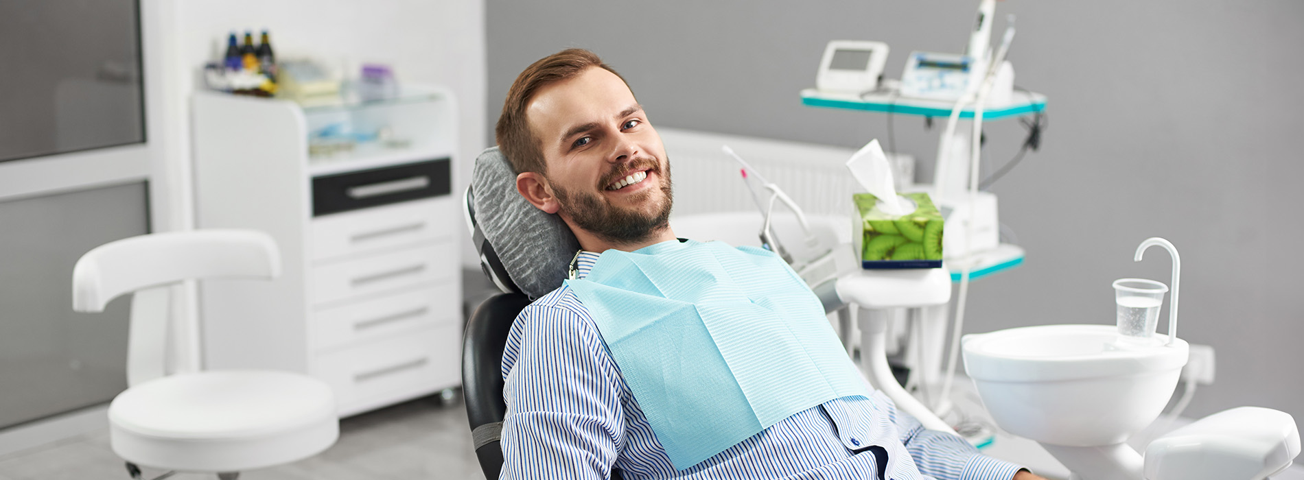 A man sitting in a dental chair with a smiling expression, wearing a blue shirt, surrounded by dental equipment.