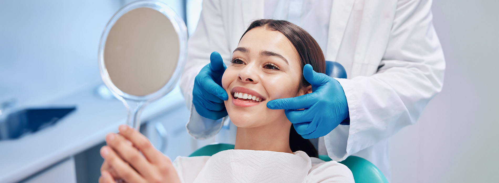 The image shows a person sitting in a dental chair with a hygienist performing an oral examination using a mirror, while another individual stands behind them wearing protective eyewear and a white coat, suggesting a professional dental care setting.