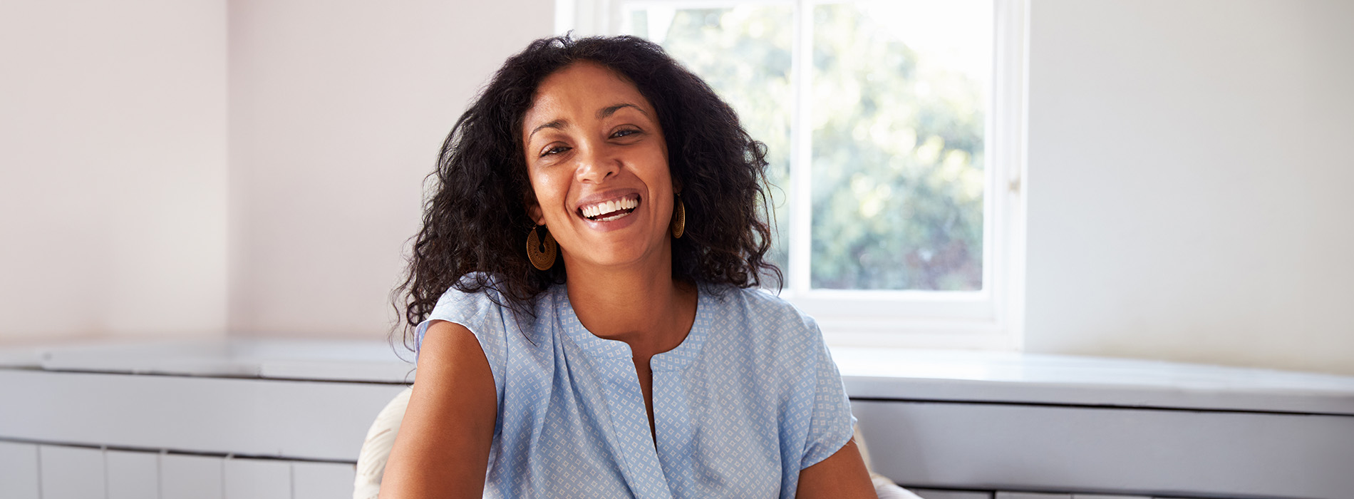 The image shows a smiling woman with curly hair sitting at a desk indoors, wearing a blue top.