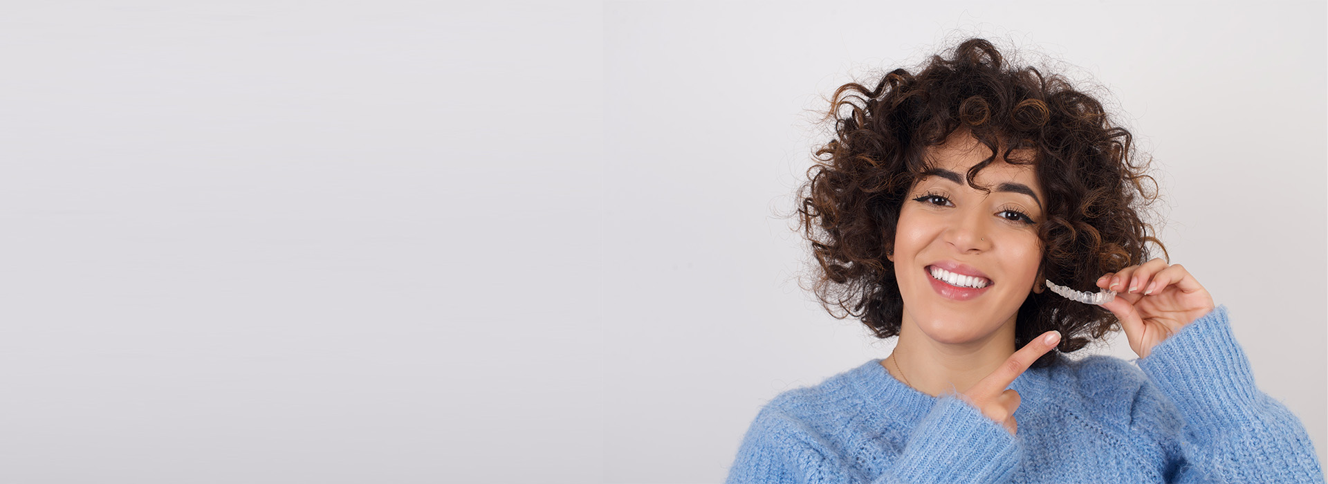 A woman with curly hair smiles and gestures towards the camera while standing against a plain background.