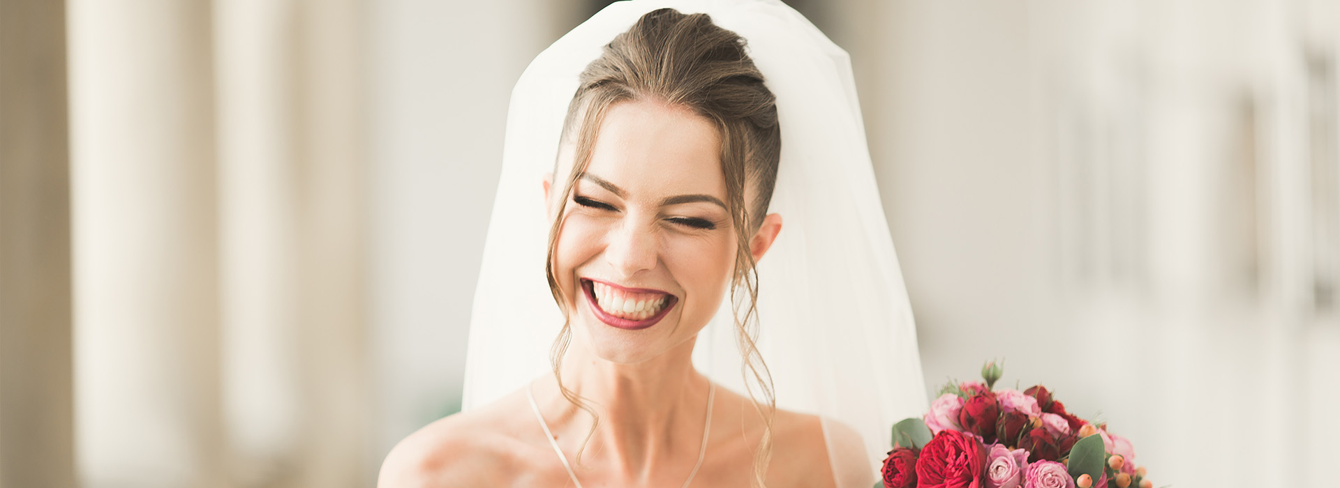 The image shows a woman wearing a wedding veil, smiling at the camera, with her hands clasped together, standing in front of a blurred background.