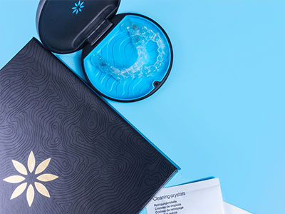 A blue dental impression tray and a toothbrush with a case are placed on a black surface next to an open booklet with a floral design, all against a light blue background.