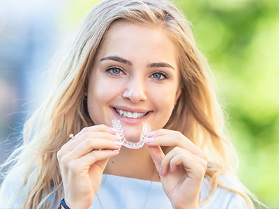 The image features a young woman with blonde hair smiling at the camera while holding up a clear plastic dental retainer.