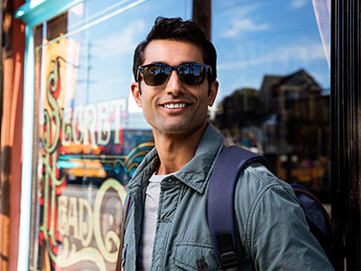 The image shows a man standing outdoors wearing sunglasses, a backpack, and a light-colored jacket, with a sign advertising  Sinbad s  visible behind him.
