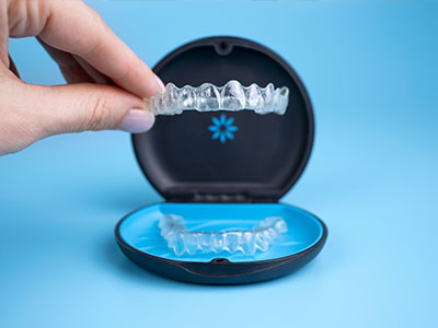 The image shows a hand holding an open case containing a clear, plastic dental retainer with a blue base.