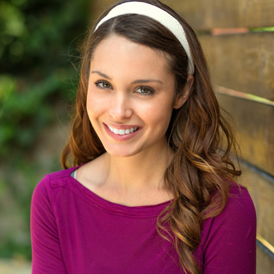 The image shows a woman with long hair wearing a purple top and a headband, smiling at the camera against a wooden fence background.