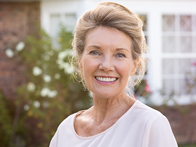 This is a portrait-style photograph of an older woman with short hair, wearing a light top, standing outdoors in front of a house with a brick wall and greenery.