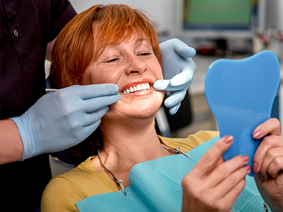 The image depicts a woman sitting in a dental chair, receiving dental care with a smile on her face, while holding up a blue toothbrush with both hands.