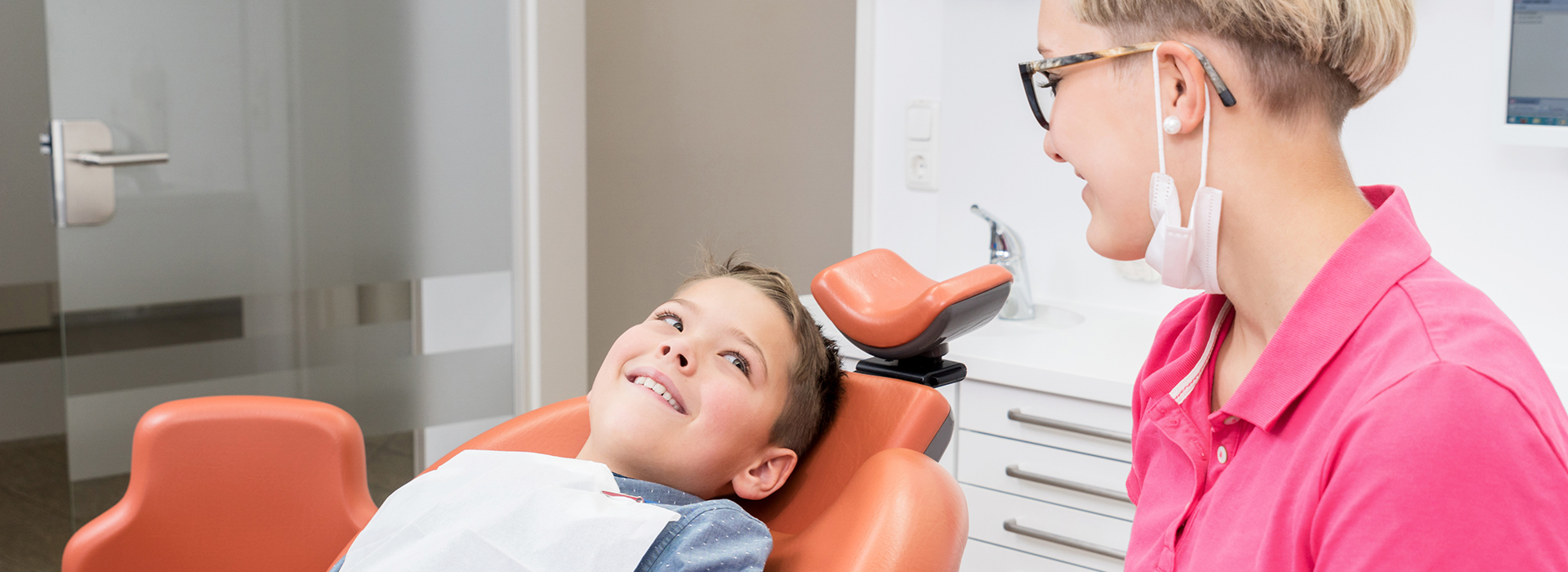 A young boy sitting in an orange dental chair, smiling at the camera, with a woman standing behind him, wearing a pink shirt, who appears to be a dentist or dental hygienist.