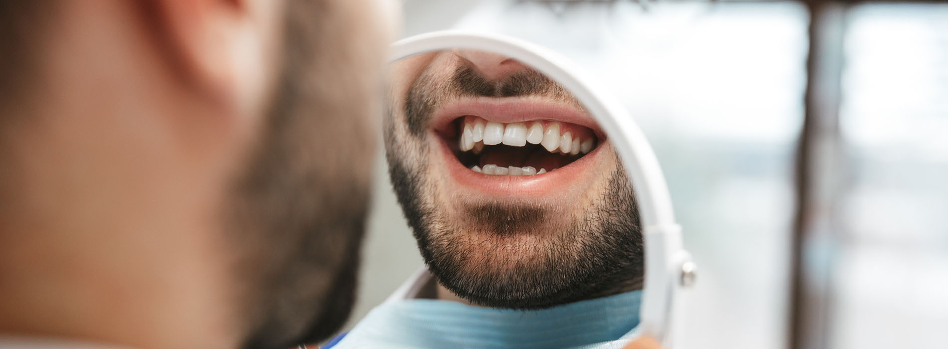 The image features a man with facial hair, wearing a blue surgical mask, sitting in a dental chair, smiling at another person who appears to be taking his picture from behind.