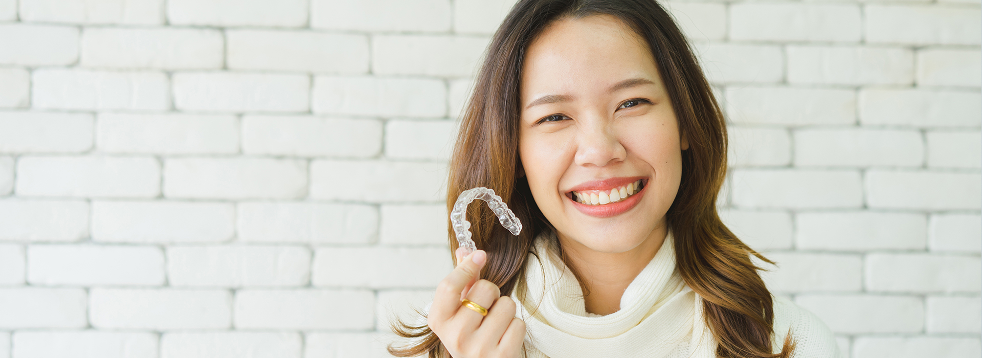The image features a smiling woman holding a ring, standing against a brick wall with a white brick pattern.