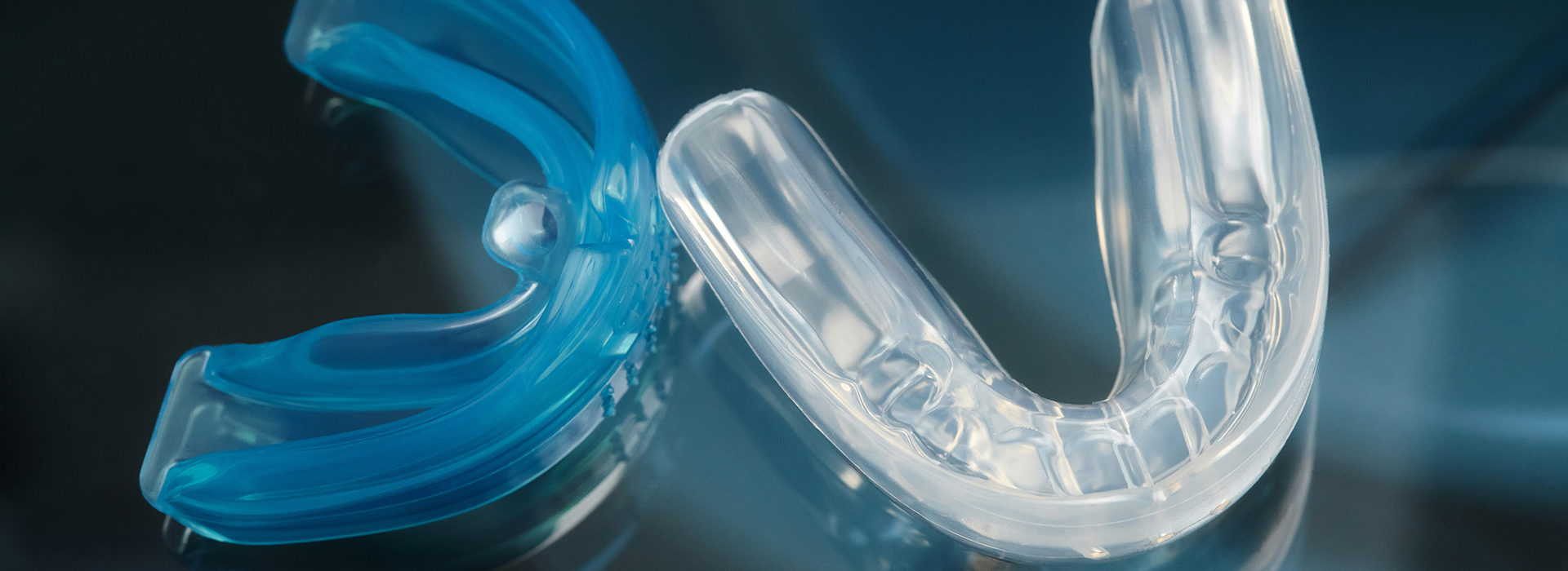 The image shows a close-up of clear, blue plastic braces with a metallic clasp, set against a blurred background that appears to be a dark surface reflecting light.