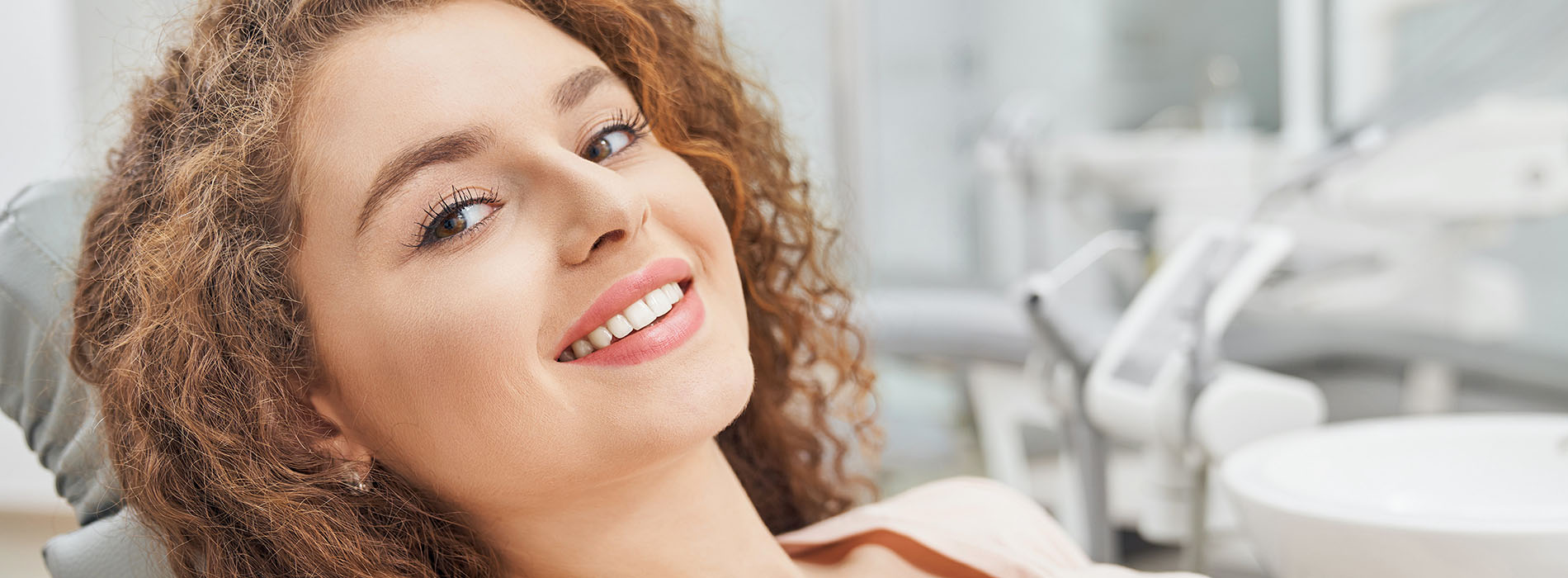 The image shows a woman with curly hair sitting in a dental chair, smiling at the camera, with her eyes closed. She appears to be relaxed or content during her dental appointment.