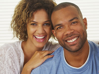 A man and woman smiling at the camera, posing together in front of a window with white blinds.