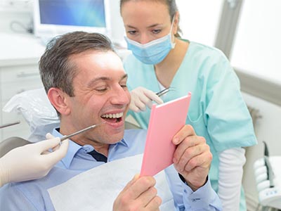 The image shows a person holding up a pink card with a smile while seated in a dental chair surrounded by dental equipment, with a dentist standing behind them.