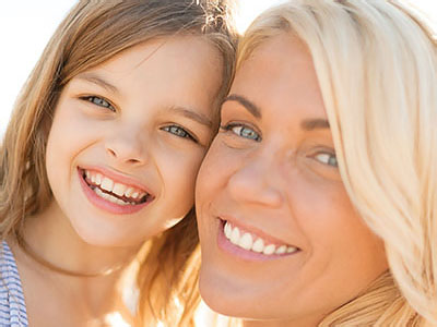 The image shows a woman with blond hair smiling at the camera, accompanied by a young girl who also smiles at the camera. They appear to be sharing a joyful moment together outdoors during daylight.