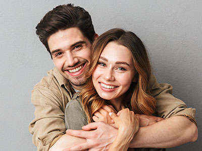 A man and woman are embracing each other with smiles, posing for a photo against a neutral background.