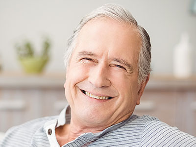 The image shows a smiling older man with gray hair, wearing a blue shirt, sitting comfortably in a chair at home.
