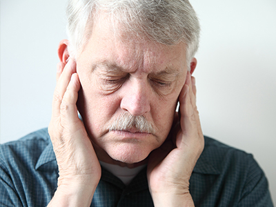 An elderly man with a mustache, wearing glasses, holds his hand to his ear, seemingly in discomfort or pain.